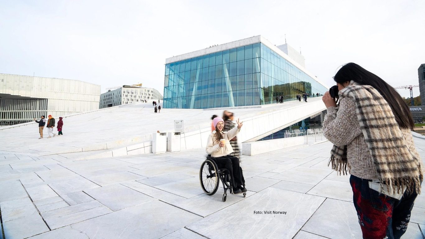 Eine Frau lächelt in die Kamera. Sie sitzt in einem Rollstuhl vor der Oper in Oslo. Foto: Visit Norway - Frau im Rollstuhl in Oslo